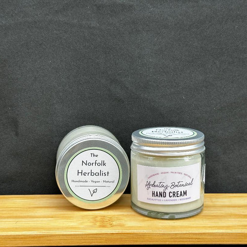 Two small round jars of The Norfolk Herbalist products on a wooden surface against a dark gray background. The left jar is labeled 'Herbalist Herbalist Hand Cream' and the right jar is labeled 'The Norfolk Herbalist' with a white lid.